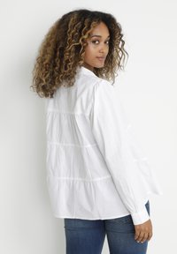 White cotton shirt featuring a loose fit, gathered seams, and button cuffs. Styled with blue jeans, showcasing a soft, smooth texture.