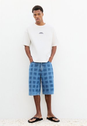 Homme debout les mains dans les poches portant un T-shirt blanc oversize, un short en jean bleu à carreaux et des tongs noires sur un sol en gravier clair.
