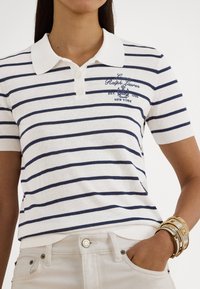 Biele polo tričko s námorníckymi vodorovnými pruhmi, krátkymi rukávmi a golierom. Vyšitý logo na ľavej strane hrudníka. Nosené s nohavicami svetlej farby a zlatými náramkami.