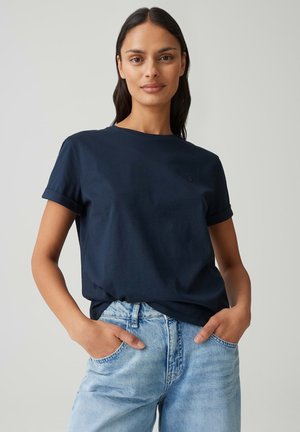 Donna con capelli lunghi e scuri che indossa una T-shirt blu navy a maniche corte infilata in un paio di jeans azzurri, mani nelle tasche, sfondo neutro.