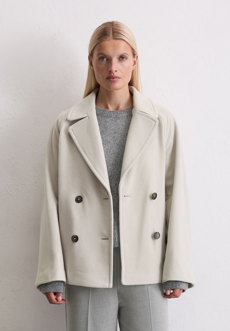 Veste en polaire légère grise à double boutonnage avec grands revers, deux boutons et un pull en tricot gris texturé en dessous. Coupe ajustée avec des manches droites.