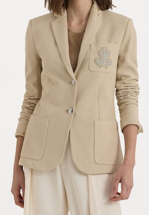 Kobieta ubrana w beżową marynarkę z dwoma guzikami i wyszytym monogramem na kieszeni na piersi, zestawioną z jasnokrępowymi szerokimi spodniami.