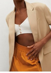 Blazer beige oversize sopra un crop top bianco a coste con bottoni, abbinato a pantaloncini arancioni con vita elastica; tessuto liscio e taglio rilassato.