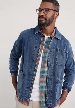 Denim jacket with a button front, two front pockets, and a collar, layered over a plaid shirt with various colors and patterns underneath.