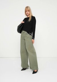 Črna majica z dolgimi rokavi z belim trakom, kombinirana s širokimi hlačami oljčno zelene barve. Model drži črno torbico in nosi črne čevlje z ostrimi konicami.