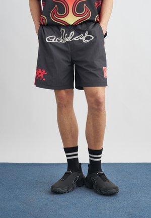 Άτομο που φοράει μαύρο σορτς με λευκό γραφικό κείμενο, μαύρες κάλτσες με λευκές ρίγες και μαύρα παπούτσια χωρίς κορδόνια, στέκεται σε μπλε χαλί.