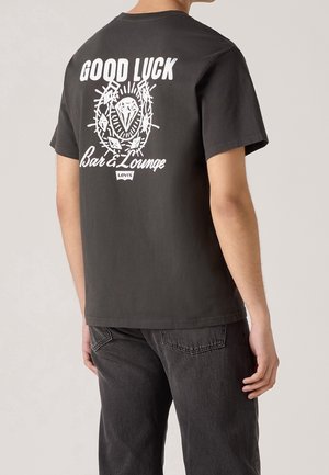 Mand iført sort Levi's T-shirt med hvid "GOOD LUCK Bar & Lounge" tekst og grafik af en diamant og torne på bagsiden.