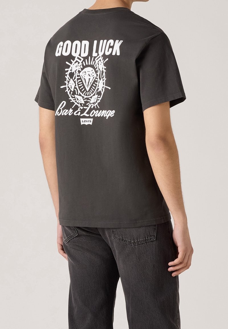 Homme portant un T-shirt noir Levi's avec un texte blanc "GOOD LUCK Bar & Lounge" et un graphique représentant un diamant et des épines dans le dos.