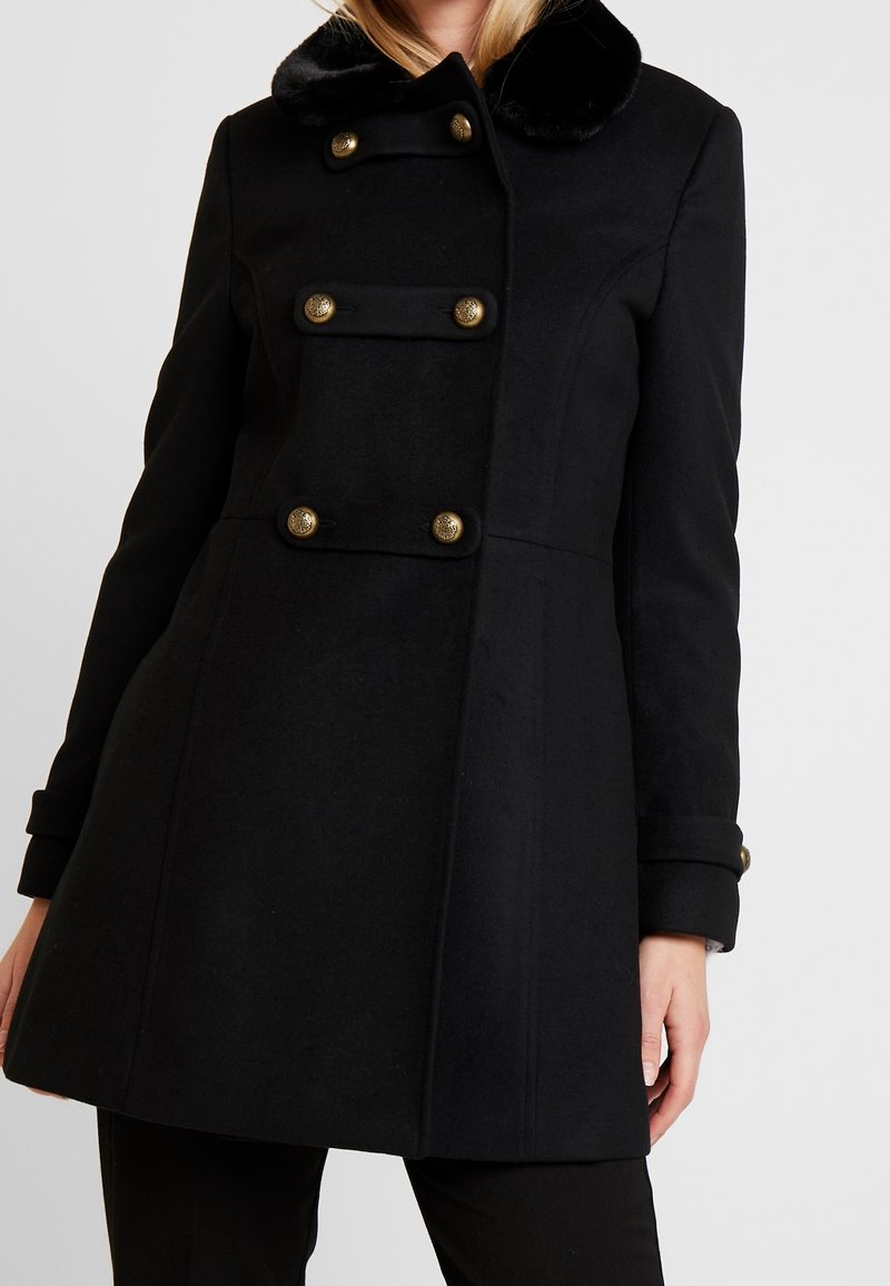 Kobieta ubrana w czarny dwurzędowy płaszcz z złotymi guzikami i czarnym futrzanym kołnierzem, stojąca na tle jednolitego jasnego tła.