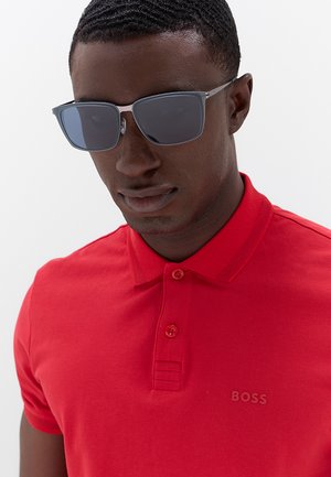 Uomo con capelli corti che indossa occhiali da sole scuri e una polo rossa BOSS su uno sfondo chiaro uniforme.