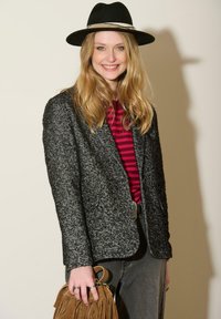 Blazer texturé noir et gris sur une chemise rayée rouge et noire, associé à un jean foncé et un sac en cuir frangé marron. Chapeau noir à large bord.