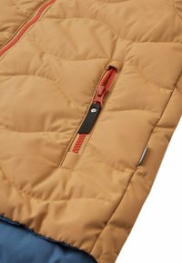 Giacca imbottita trapuntata di color marrone chiaro con cerniere rosse. Presenta una tasca laterale con chiusura a scatto nera. Texture morbida e liscia.