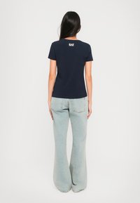 Mørkeblå bomulls T-skjorte med korte ermer, tettsittende design, har hvit EA7-logo på baksiden; kombinert med lyseblå flared jeans.