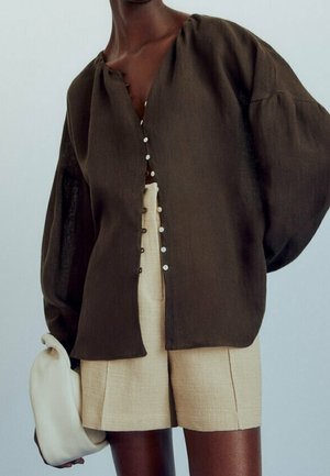 Persoon draagt een donkerbruine blouse met knopen en volumineuze mouwen, beige textuurrijke korte broek en houdt een zachte witte clutch vast.