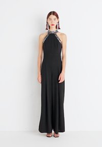 Černé dlouhé halter šaty s výšivkou v oblasti výstřihu, skládáním a minimalistickým designem. Modelka nosí velké náušnice.