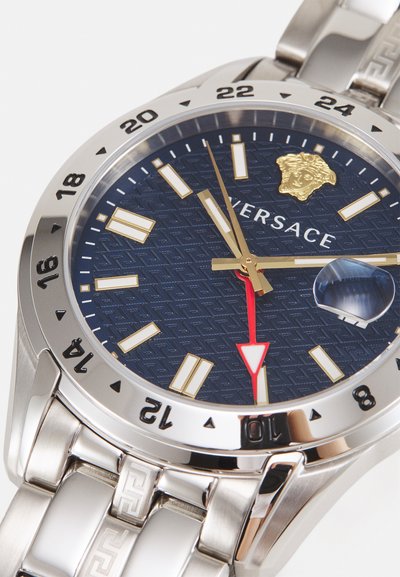 Montre bracelet en argent avec un cadran texturé bleu, des index horaires dorés, une aiguille des secondes rouge et un logo Méduse doré à 12 heures.