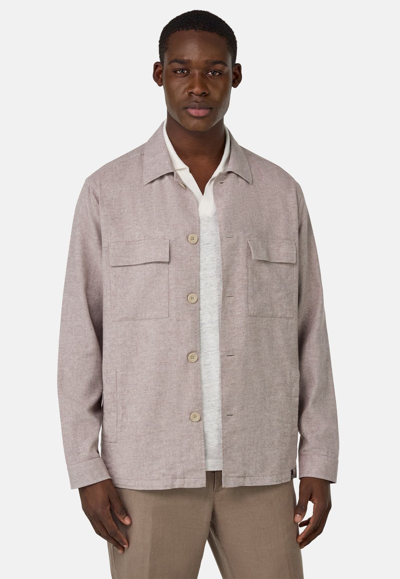 Chaqueta beige clara hecha de tejido texturizado, con un cuello con solapa, dos bolsillos en el pecho y cierre de botones, llevada sobre una camisa gris.