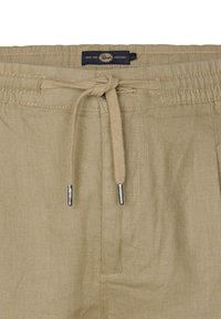 Pantalon en lin beige avec taille élastique et cordon de serrage, doté d'embouts en métal argenté et d'une petite étiquette marine avec le texte de la marque.