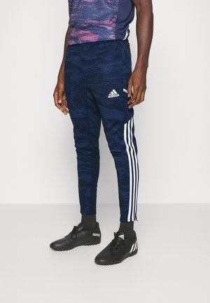 Homme portant un pantalon de sport Adidas bleu marine avec des bandes blanches et des chaussures de sport Adidas noires, debout devant un fond uni.