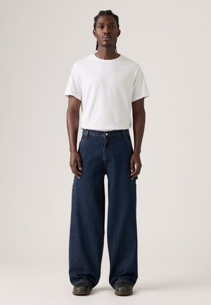 Homme debout portant un t-shirt blanc uni, un jean large bleu foncé et des chaussures noires sur un fond neutre.