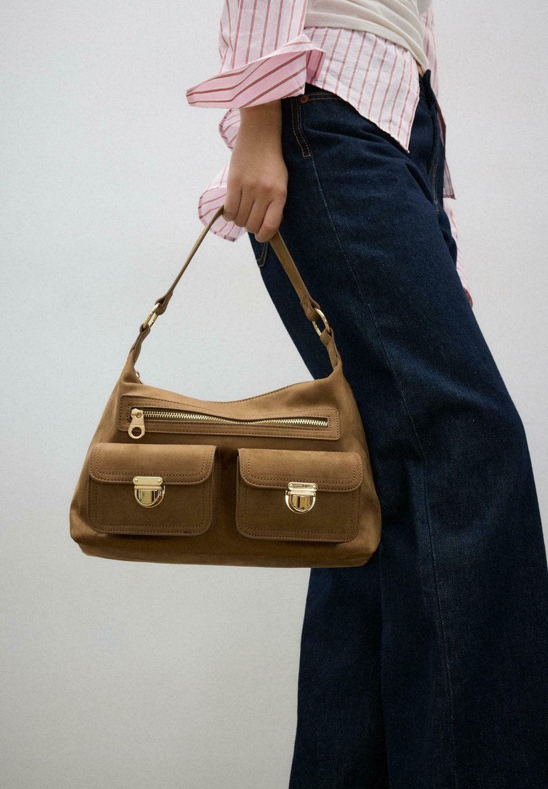 Personne tenant un sac à main en daim marron avec une fermeture éclair et deux poches avant, portant un jean foncé et une chemise rayée.