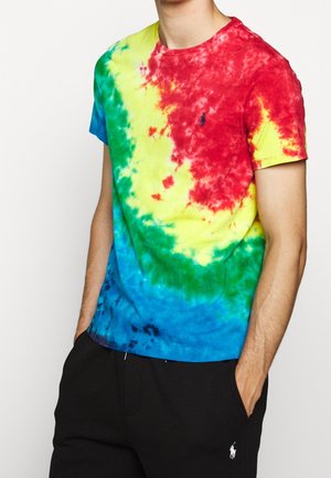 Homme portant un t-shirt tie-dye à manches courtes rouge, jaune, vert et bleu avec un pantalon noir à poches, mains partiellement dedans.