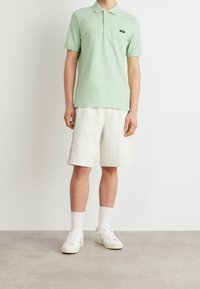 Homme portant un polo vert clair à manches courtes, un short blanc longueur genou avec bande latérale, des chaussettes blanches et des baskets blanches, debout sur un sol gris.