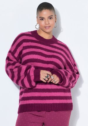Ung kvinde med kort hår iført hoop-øreringe, en pink og lilla stribet sweater og mørkerøde bukser, der poserer med hænderne foldet.