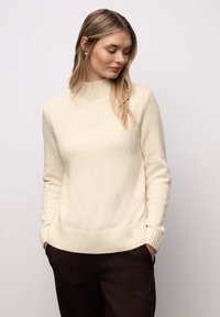 Cremefarbener Rollkragenpullover aus weichem Strickmaterial, mit gerippten Bündchen und Saum, kombiniert mit dunkelbraunen Hosen.