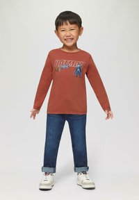 Langarmige roestkleurige shirt met een Batman-afbeelding, gecombineerd met opgerolde blauwe denim jeans en witte sneakers met grijze accenten.