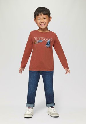 Langarmige roestkleurige shirt met een Batman-afbeelding, gecombineerd met opgerolde blauwe denim jeans en witte sneakers met grijze accenten.