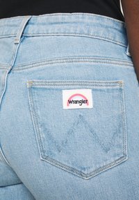 Hellblaue Denim-Jeans mit einem weißen "Wrangler"-Label, das einen rosa Regenbogen zeigt. Auffälliges, gesticktes "W"-Muster auf den Gesäßtaschen.