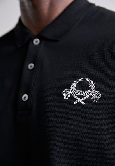 Svart polo-skjorte laget av teksturert stoff med en tre-knappsklaff, som har en hvit brodert Moschino-logo over kryssede symboler.