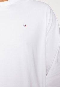 Hvit langermet T-skjorte laget av myk bomull. Har en liten, brodert logo i rød, blå og hvit på venstre brystområde.