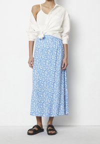 Modrá květinová maxi sukne s bílým vzorem, kombinovaná se světlejší bílou košilí se zavázaným uzlem a černými sandály. Hladká tkanina, splývavý design.