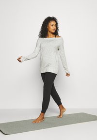 Maglietta grigia chiara a maniche lunghe con design sceso da spalle e fori per i pollici, abbinata a pantaloni jogger neri, mentre cammina su un tappetino da yoga grigio.