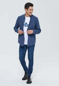 Chemise bleue à boutons portée sur un t-shirt blanc avec un graphisme bleu, associée à un jean en denim foncé et des bottes noires à lacets. Style décontracté.