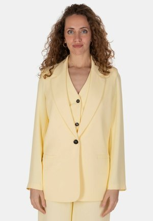 Donna con capelli ricci che indossa un blazer giallo pallido con bottoni neri, in piedi davanti a uno sfondo chiaro uniforme, rivolta in avanti.