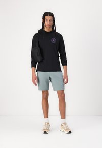 Haut à capuche noir à manches longues avec logo, shorts verts clairs et chaussures de sport de couleur neutre. Petit sac en bandoulière noir porté sur le côté gauche.