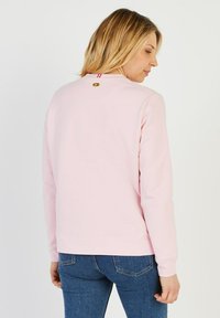 Sweatshirt rose clair avec un col rond, des poignets et un ourlet côtelés. Présente un détail de logo doré à l'arrière du col et une texture lisse.