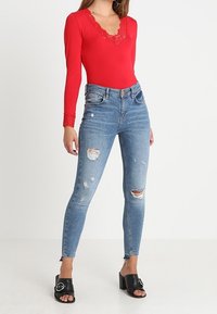 Body a maniche lunghe rosso con bordo in pizzo, abbinato a jeans skinny blu a vita alta strappati e sandali neri a ciabatta con dettagli in metallo.