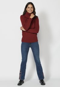 Rdeča pletena turtleneck pulover z rebrasto teksturo, v kombinaciji z modrimi kavbojkami z cvetličnimi akcenti na robu in črnimi gležnarji.