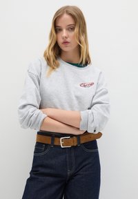 Giovane donna dai capelli biondi si trova con le braccia incrociate, indossando una felpa grigia con un logo rosso e jeans blu scuro con una cintura marrone.