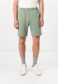 Shorts en coton verts avec une taille élastique, des poches latérales et un détail de logo. Portés avec des chaussettes grises et des baskets blanches.