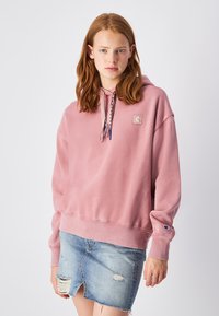 Sudadera de algodón rosa con un parche de logo en el frente, cuerditas con acentos en azul y beige, combinada con una falda de mezclilla desgastada.