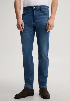 Mann trägt blaue Slim-Fit-Jeans und dunkelbraune Chelsea-Stiefel aus Wildleder und steht vor einem schlichten hellen Hintergrund.