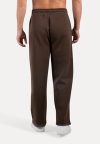 Mann in lockeren braunen Jogginghosen und weißen Sneakers, von hinten gezeigt, vor einem schlichten Hintergrund stehend.