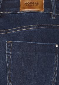Jeans de mezclilla azul oscuro con un parche de cuero etiquetado "MORGAN" y un bolsillo trasero con costuras visibles y un acento metálico decorativo.