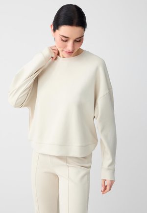 Donna con capelli scuri che indossa una felpa crema a maniche lunghe e pantaloni abbinati, guarda verso il basso con una mano vicino al collo.