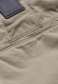 Beige stof met een gladde textuur, voorzien van zichtbare stiksels en een zwart rechthoekig label met het cijfer "5" dat aan de bovenkant is bevestigd.